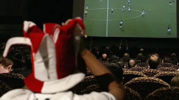 Cinema attendees sitting in patterned theatre seats watching a football match on a large screen, with a fan in a red and white jester hat in the foreground.