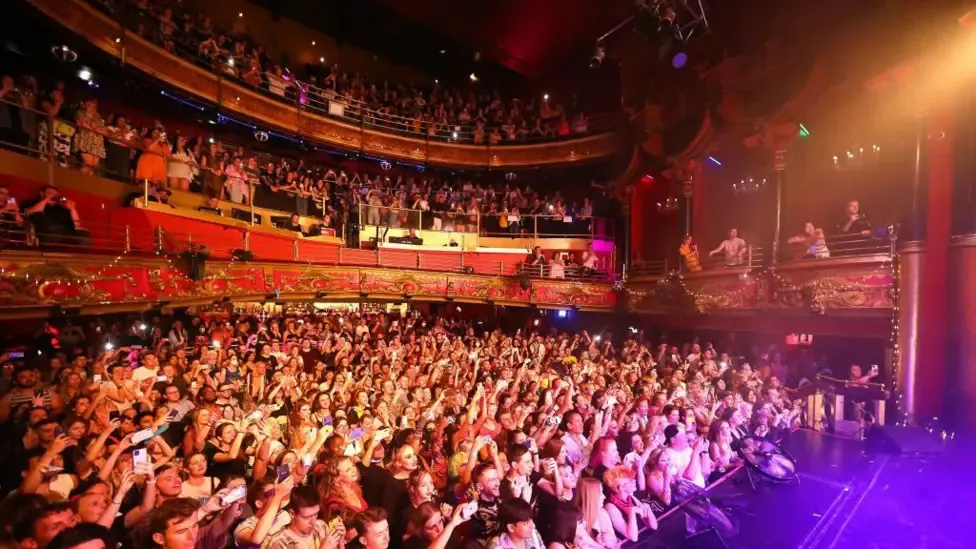 A massive, dense crowd of people with upraised hands packing the floor and balconies of a historic, red-lit music hall venue.