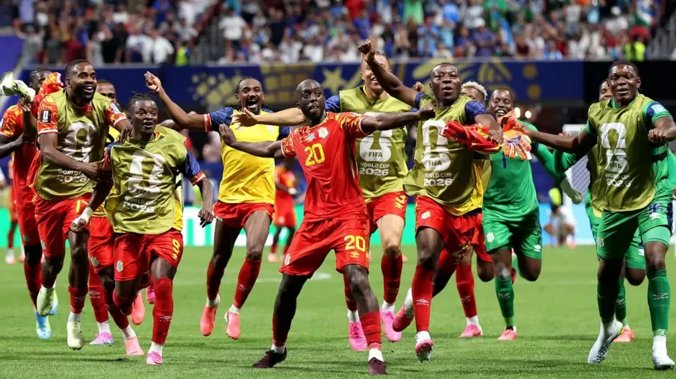 Yoane Wissa spreads his arms out as he dances in celebration after scoring a goal against Uzbekistan and the World Cup finals. He is surrounded by several DR Congo substitutes, who all bear smiles as some of them copy his pose